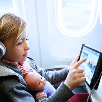 小女孩戴着耳机，腿上放着一个娃娃，在飞机上的WiFi上用平板电脑玩游戏